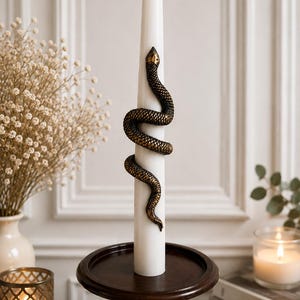 May include: A tall white taper candle featuring a textured, dark bronze-colored snake coiled around its center. The snake has detailed scales and a defined head pointing upward. The candle is positioned upright on a dark brown wooden candle holder.