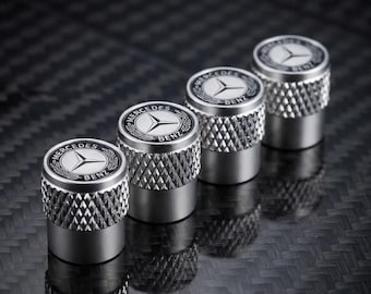 Puede incluir: Cuatro tapones de válvula de neumático de metal plateado con textura moleteada. Cada tapón presenta un emblema circular en blanco y negro en la parte superior con el logotipo y texto de Mercedes-Benz, dispuestos en diagonal sobre fibra de carbono.
