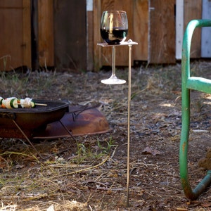 May include: A metal wine glass holder with a long metal stake. The holder is designed to hold a wine glass upright. The holder is in a grassy area with a grill in the background.