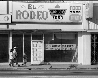 May include: A black and white photo of a storefront with a sign that reads "El Rodeo Mexican Food" and "Food To Go Tortilleria". The storefront has a large window with a sign that reads "Restaurant For Rent (213) 464-5006 464-4905".