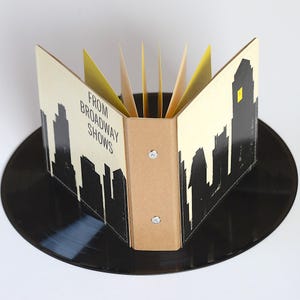 May include: A small binder with a cream-colored cover featuring a black silhouette of a city skyline and the text "FROM BROADWAY SHOWS" in black lettering. The binder is open, revealing yellow interior pages, and is resting on a black vinyl record.