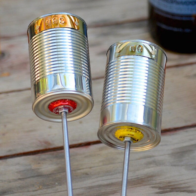 Puede incluir: Dos adornos de jard&iacute;n hechos a mano con latas recicladas. Las latas plateadas tienen bordes dorados y est&aacute;n unidas a varillas met&aacute;licas. Una tiene una tapa de botella roja, la otra amarilla. El fondo es de madera.
