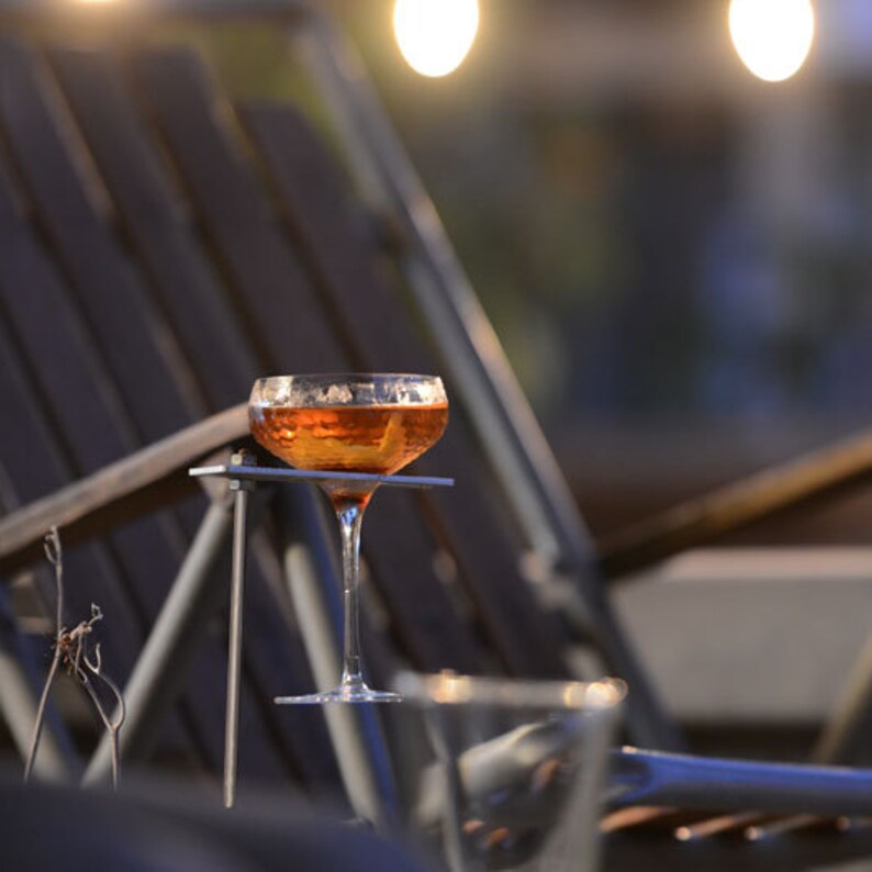 May include: A clear glass cocktail glass with a gold rim and a brown alcoholic beverage inside. The glass is sitting on a metal stand that is attached to a wooden chair.