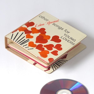 May include: A vintage CD case with the text "other Special songs for YOUNG LOVERS" and red and orange heart illustrations. The case is beige with a brown spine and contains several CDs. A CD is visible at the bottom.