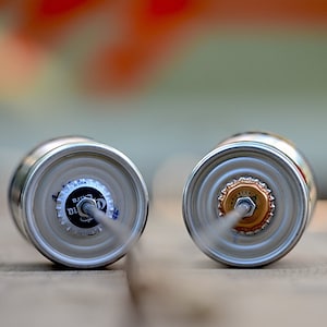 Puede incluir: Dos latas de cerveza plateadas con sus tapas mirando al espectador. Una tapa es blanca y negra con el texto "Corona Extra". La otra tapa es dorada. Ambas latas tienen una pajita insertada.