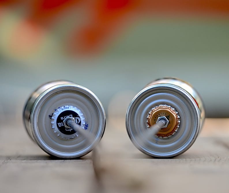 Puede incluir: Dos latas de cerveza plateadas con sus tapas mirando al espectador. Una tapa es blanca y negra con el texto "Corona Extra". La otra tapa es dorada. Ambas latas tienen una pajita insertada.
