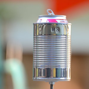 Puede incluir: Una lata de metal reutilizada con una tapa de leng&uuml;eta, transformada en un objeto decorativo. El exterior plateado de la lata es acanalado y la parte superior presenta detalles en rosa y blanco. Las iniciales "MR" est&aacute;n estampadas en el lateral.