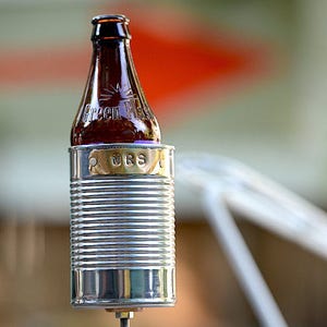 Puede incluir: Una botella de vidrio marr&oacute;n con el texto "Green River" est&aacute; colocada en una lata de metal plateado. Una peque&ntilde;a placa dorada con las letras "MRS" est&aacute; adherida a la lata. La lata tiene un dise&ntilde;o acanalado.