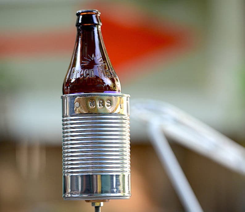 Puede incluir: Una botella de vidrio marr&oacute;n con el texto "Green River" est&aacute; colocada en una lata de metal plateado. Una peque&ntilde;a placa dorada con las letras "MRS" est&aacute; adherida a la lata. La lata tiene un dise&ntilde;o acanalado.