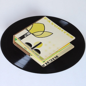 May include: A square, cream-colored notebook with the word "FAVORITES" in gold lettering and a yellow floral design. The notebook is resting on a black vinyl record. The notebook has a brown border and colorful pages.