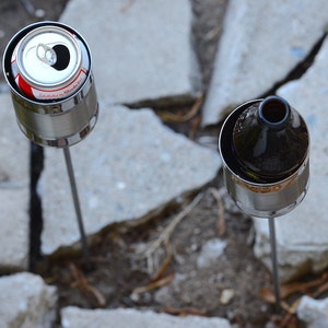 Puede incluir: Dos antorchas de jard&iacute;n caseras hechas de latas recicladas. Una contiene una lata de bebida abierta, la otra una botella de vidrio marr&oacute;n. Ambas est&aacute;n montadas sobre estacas de metal, sobre un fondo de adoquines de hormig&oacute;n y cobertura vegetal natural.