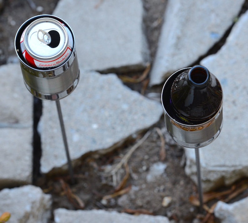 Puede incluir: Dos antorchas de jard&iacute;n caseras hechas de latas recicladas. Una contiene una lata de bebida abierta, la otra una botella de vidrio marr&oacute;n. Ambas est&aacute;n montadas sobre estacas de metal, sobre un fondo de adoquines de hormig&oacute;n y cobertura vegetal natural.