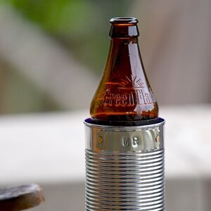 Puede incluir: Una botella de vidrio marr&oacute;n con el texto "Green Flash" se encuentra en un enfriador de latas plateado. El enfriador tiene un dise&ntilde;o acanalado y las iniciales "MB" son visibles. El fondo est&aacute; borroso.