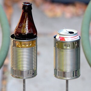 Puede incluir: Dos soportes met&aacute;licos para latas, uno con una botella de vidrio marr&oacute;n con el texto "Green Flash", y el otro con una lata blanca y roja. Ambos soportes est&aacute;n sujetos a varillas met&aacute;licas. Los soportes tienen una banda dorada con las letras "MBG" y "MR".