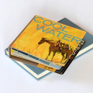 May include: A square, yellow-toned book cover featuring the text COOL WATER in blue letters above a dark illustration of a horse with a saddle. The cover is stitched along the edges and rests on top of a larger, plain blue hardcover book.