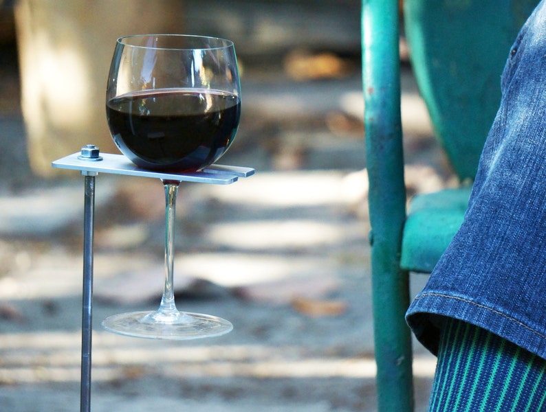 May include: A metal wine glass holder with a silver finish. The holder is attached to a metal rod that is inserted into the ground. A glass of red wine is resting in the holder.