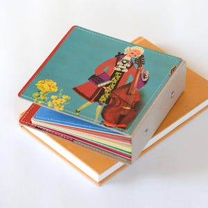 May include: A stack of colorful notebooks with a decorative cover featuring a cartoon illustration of a person in a red coat playing a cello. The cover has a teal background with yellow flowers.
