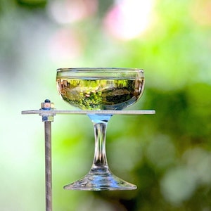 May include: A clear glass champagne flute filled with a pale yellow liquid sits on a silver metal stand. The stand has a single metal rod extending from the base.