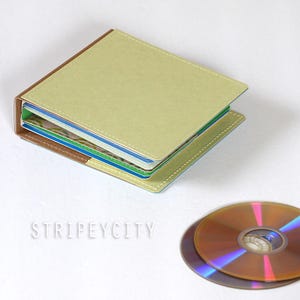May include: A square, light green CD case with brown spine accents and white stitching along the edges. The case is open, revealing internal sleeves for discs. A single, reflective, iridescent CD is positioned next to the case. The text STRIPEYCITY is visible.