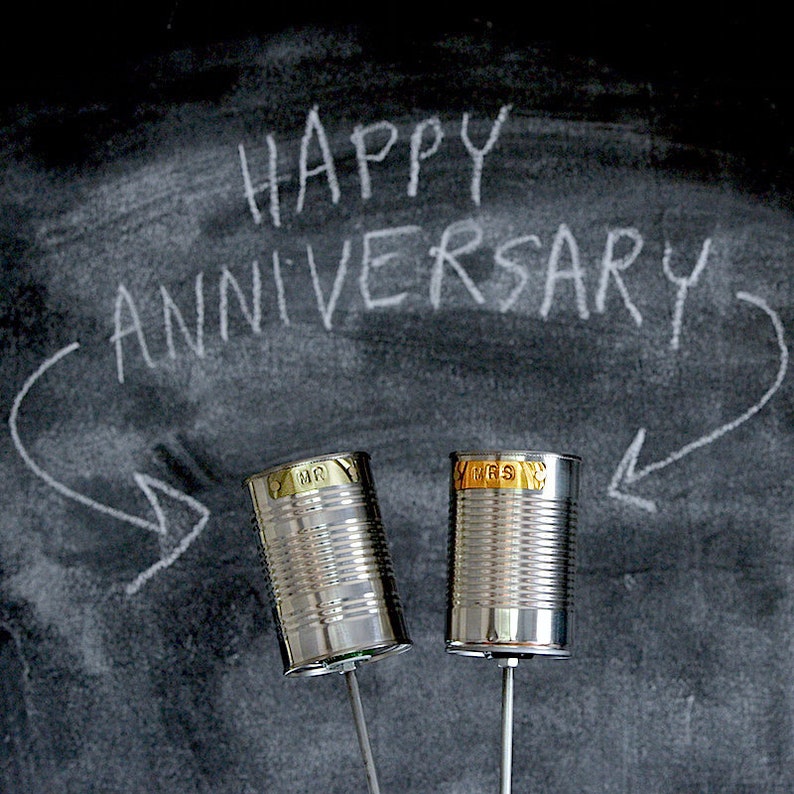 Puede incluir: Dos decoraciones de latas de conserva plateadas con las palabras "MR" y "MRS", montadas sobre varillas met&aacute;licas. El fondo es una pizarra con las palabras "HAPPY ANNIVERSARY" escritas con tiza blanca, con flechas.