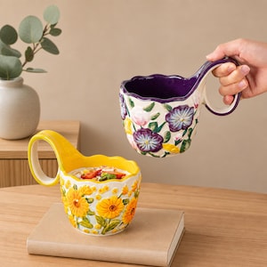 Può includere: Due tazze in ceramica con bordi smerlati e grandi manici. Una tazza è gialla con girasoli gialli in rilievo e foglie verdi. L'altra tazza è viola all'interno e bianca all'esterno con motivi floreali viola e rosa in rilievo.