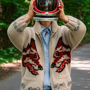 May include: A beige knit zip-up cardigan featuring a red and black fox pattern on the front, a shawl collar, and ribbed cuffs. The garment is worn over a light blue button-down shirt and blue jeans, with a red astronaut helmet held overhead.