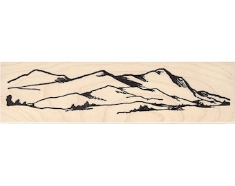 May include: Black and white line drawing of a mountain range with a few trees in the foreground. The image is on a light brown wooden background.