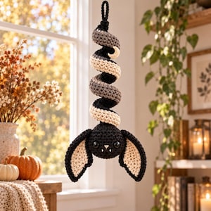 May include: A crocheted hanging ornament featuring a black bat with large ears and a spiral tail in black, grey, and cream. The tail includes a small cream-colored crescent moon accent. The bat has two black eyes and two small white fangs.