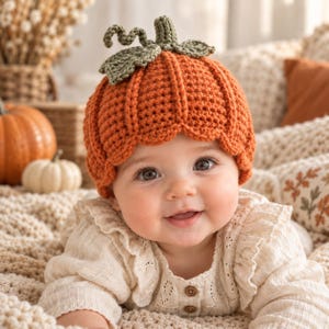 Puede incluir: Un gorro de calabaza tejido a ganchillo en color naranja vibrante, con un diseño acanalado texturizado y un tallo verde tejido con dos hojas en la parte superior. El gorro tiene un borde festoneado y está hecho con hilo grueso.