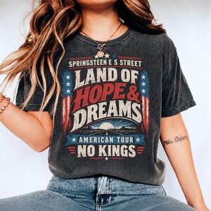 May include: A charcoal grey short-sleeve t-shirt featuring a distressed graphic print with the text "SPRINGSTEEN E S STREET LAND OF HOPE & DREAMS AMERICAN TOUR NO KINGS". The design includes red, white, and blue flag motifs and a road scene.