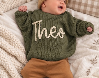 Puede incluir: Un bebé con un suéter de punto verde oliva de manga larga con el nombre Theo bordado en hilo color crema en el pecho, combinado con un gorro color canela y pantalones marrones lisos. El suéter presenta una textura acanalada y cuello redondo.