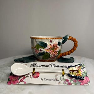 May include: A ceramic teacup with a floral design and a braided handle, resting on a saucer. In front, a white ceramic spoon with a butterfly handle is attached to a card labeled Botanical Collection By CornerRuby with floral prints.