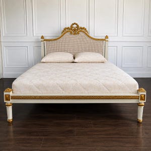 May include: A bed frame featuring a cream-coloured finish with gold-toned accents on the headboard and footboard. The headboard includes a woven cane panel and a decorative carved crest. Two cream-coloured pillows rest on a quilted cream mattress.