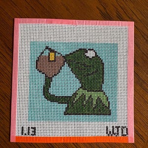 May include: A square needlepoint canvas featuring a green frog character drinking from a brown mug against a light blue background. The design is framed by a pink border with an orange stripe at the bottom and black text in the lower corners.