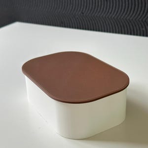 Pode incluir: Um recipiente de armazenamento retangular com cantos arredondados, apresentando uma base branca sólida e uma tampa plana castanha texturizada. O recipiente tem um acabamento mate liso nas laterais e uma superfície superior ligeiramente côncava.