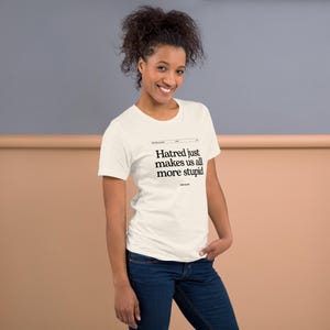 May include: A cream-coloured short-sleeve t-shirt featuring black text printed on the chest that reads: Hatred just makes us all more stupid, with the name John Hughes printed in a smaller font size directly below the main quote.