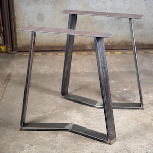 Può includere: Una coppia di gambe per tavolo in stile industriale realizzate in acciaio grezzo grigio scuro. Ogni gamba presenta una piastra di montaggio rettangolare piatta sulla parte superiore con fori preforati, collegata a una base a V da due barre di supporto angolate.