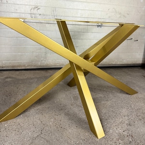 Puede incluir: Una base de mesa de metal de color dorado con un diseño geométrico en forma de X con cuatro patas anguladas que se cruzan en un punto central. La estructura incluye un marco superior rectangular con placas de montaje.