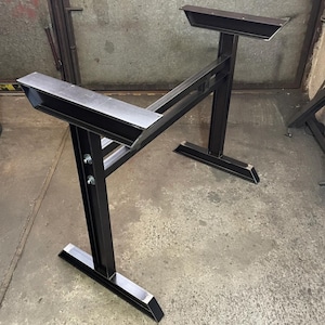 May include: A black metal table base featuring two vertical support columns connected by a horizontal crossbar. The structure includes flat mounting plates at the top and wide, stable feet at the bottom, secured with visible silver bolts and nuts.