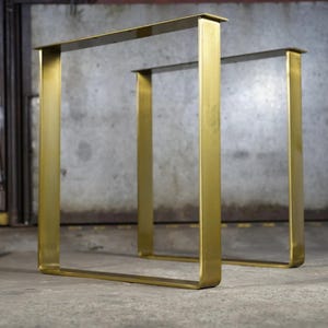 May include: Two U-shaped table legs made of brushed gold-toned metal. Each leg features a flat horizontal top mounting plate with pre-drilled holes for attachment, connected to two vertical side bars and a single horizontal base bar.