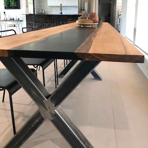 Può includere: Un lungo tavolo da pranzo caratterizzato da una superficie in legno naturale marrone chiaro con un ampio e liscio inserto centrale in resina nera opaca. Il tavolo è sostenuto da una robusta base in metallo a forma di X in acciaio grigio scuro.