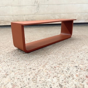 May include: A rectangular, open-ended shelf with a matte, terracotta-colored finish. The shelf features a continuous, bent metal construction with rounded corners on the bottom edges and a flat, horizontal top surface for storage or display.