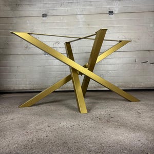 May include: A gold-coloured metal table base featuring a geometric design with four angled legs that intersect at a central point. The structure includes a rectangular frame at the top for supporting a tabletop, constructed from smooth, solid metal bars.