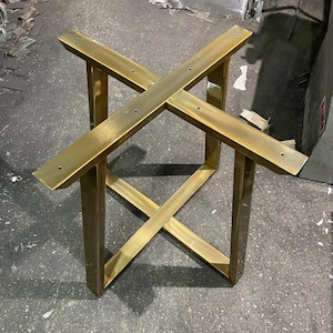 Könnte beinhalten: Ein goldfarbenes Tischgestell aus Metall mit kreuzförmigem Design, bestehend aus vier vertikalen Beinen, die durch einen zentralen X-Rahmen verbunden sind. Die oberen horizontalen Stangen haben vorgebohrte Löcher zur Montage einer Tischplatte.