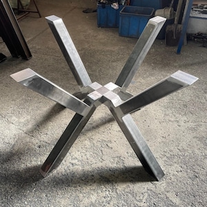 May include: A silver-colored metal table base featuring four angled legs that extend outward from a central square junction. The structure is constructed from rectangular steel tubing with a visible metallic finish and clean, sharp edges.