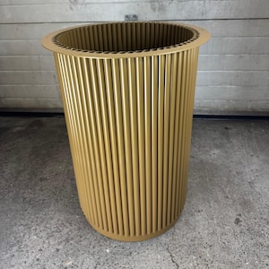 May include: A cylindrical container featuring a metallic gold finish, constructed from a series of vertical, hollow tubes arranged in a circular pattern. The top edge has a flat, circular rim with small, evenly spaced holes around the perimeter.