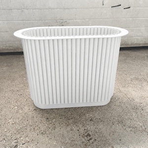 Peut inclure: Une jardinière ovale blanche dotée d'un extérieur nervuré vertical composé de nombreux tubes cylindriques fins et parallèles. Le récipient présente un rebord large et plat sur le dessus ainsi qu'une base solide et lisse, offrant un aspect uniforme.