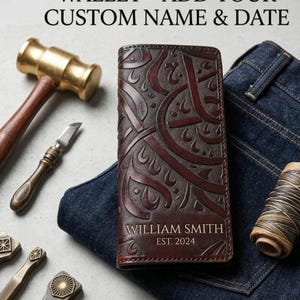 Può includere: Un portafoglio in pelle marrone scuro lavorato a mano con motivi in rilievo e il testo WILLIAM SMITH EST. 2024. Il portafoglio è circondato da strumenti in metallo, un martello di legno e un rocchetto di filo su un tessuto denim scuro.