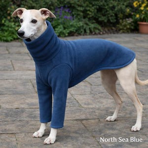 May include: A light-colored dog wearing a navy blue fleece dog coat with a high, turtleneck collar. The coat covers the dog's body and legs. The text "North Sea Blue" is visible at the bottom of the image.