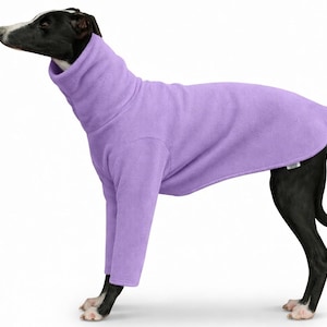 May include: A lavender dog sweater with a high turtleneck collar. The dog is black and white, wearing a fleece sweater. The sweater has long sleeves and covers the dog's body. The dog is standing on a white surface.